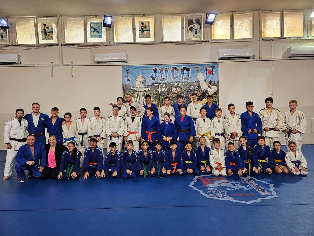 תמונה קבוצתית של ג׳ודוקאים ומאמנים על המזרן מול כרזת פסטיבל ג׳ודו בחיפה