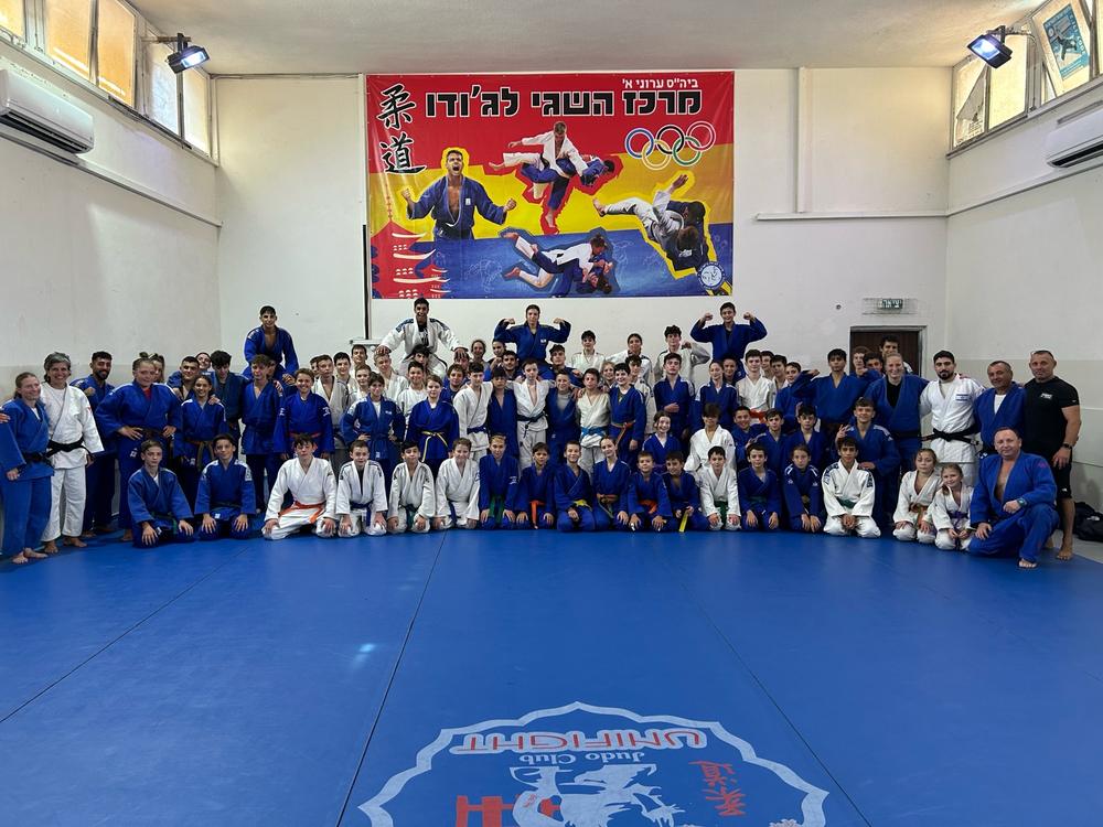 קבוצה גדולה של ג׳ודוקאים ומאמנים באולם אימונים, מתחת לכרזת ג׳ודו